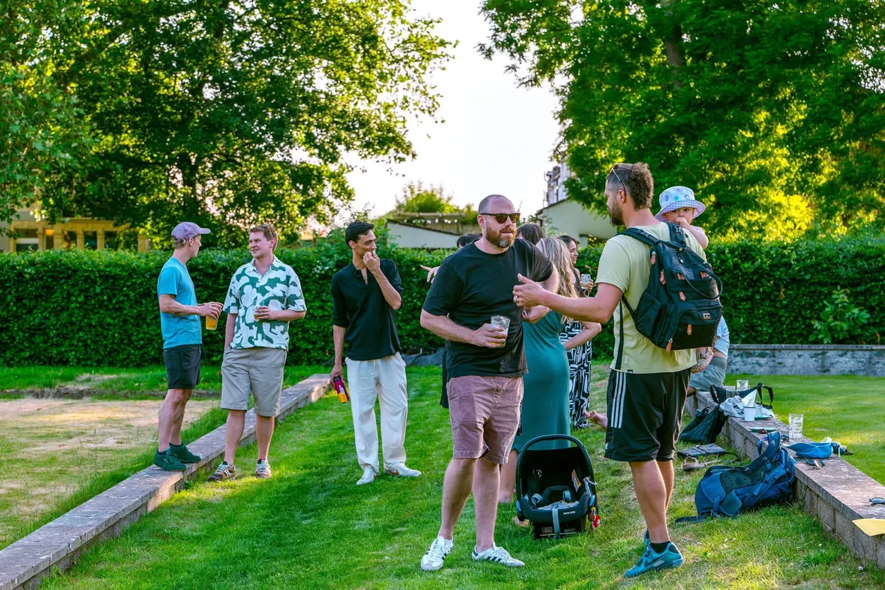 A group of people socialising on a grassy lawn with hedges and buildings in the background, some holding drinks and wearing caps.