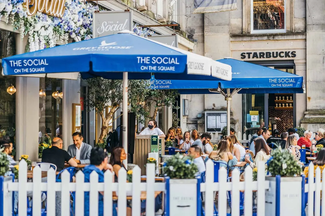 Outdoor seating area with blue umbrellas and white fencing, diners seated at tables under cover, urban square setting.