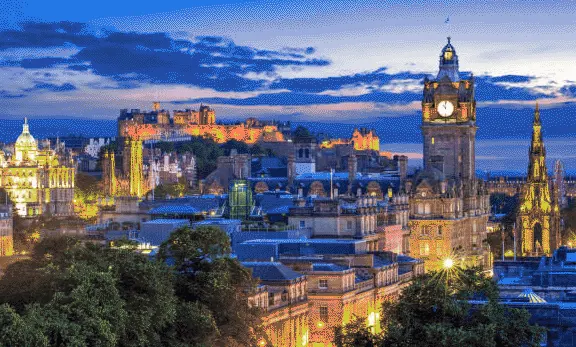 Edinburgh cityscape at dusk with illuminated historic buildings, castle and church spires under a blue twilight sky.