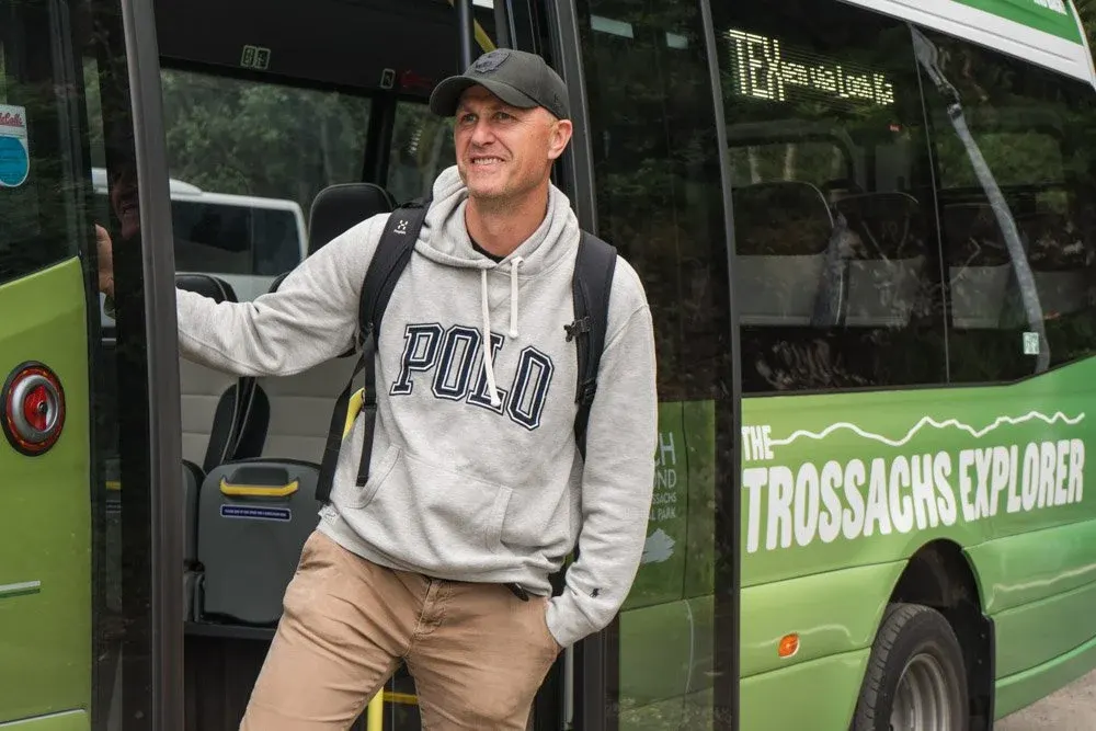 A man in casual clothing boards a green tour bus branded "Trossachs Explorer".