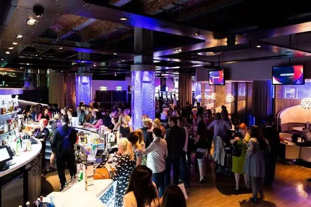 Crowded indoor venue with people socialising at bars and standing throughout a large room lit with blue and purple lighting.