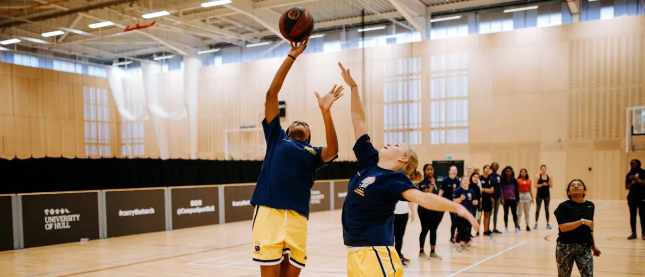 Young basketball player shooting a ball in an indoor sports hall whilst spectators watch from the sidelines.