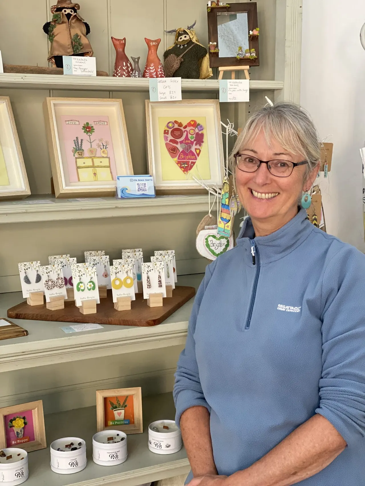 Woman with grey hair and glasses smiling beside a display of handmade jewellery and artwork in a gallery studio.