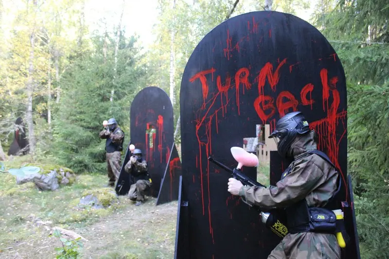 Players in protective gear and masks aim paintball guns at large black targets with red text in a wooded arena.
