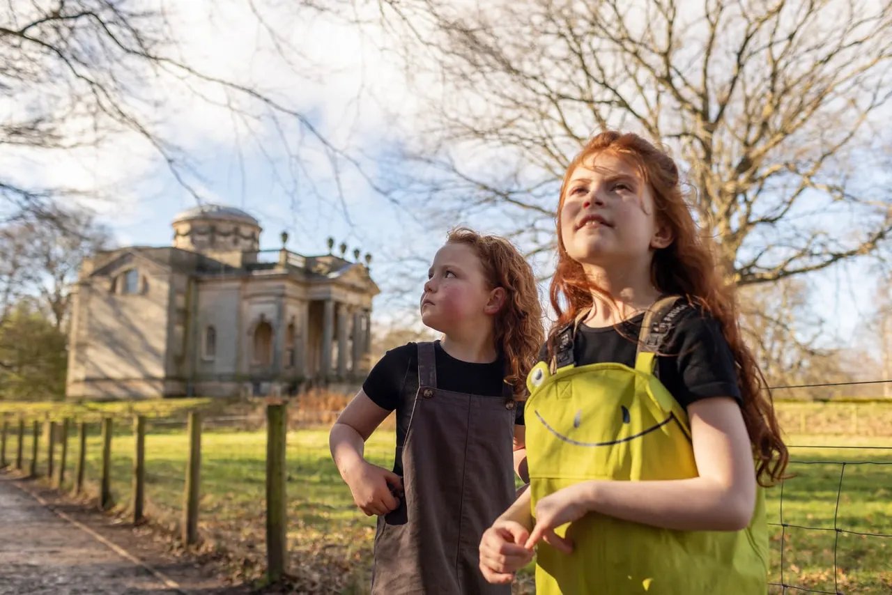 Two children stand in a countryside setting with a historic stone building and bare trees in the background.
