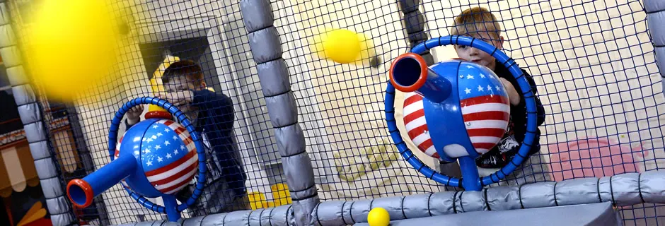 Two children playing with blue and red inflatable toys inside a padded play structure with netting.