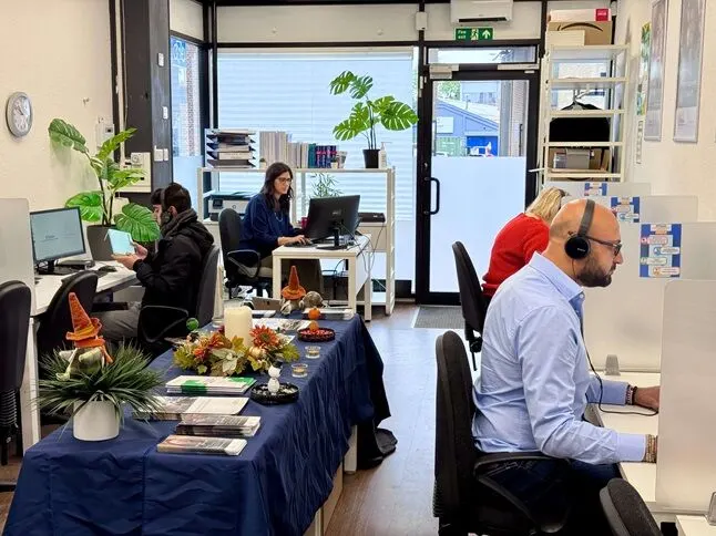 Three people working at computers in a modern training office with plants, desks and a blue-clothed table in the foreground.