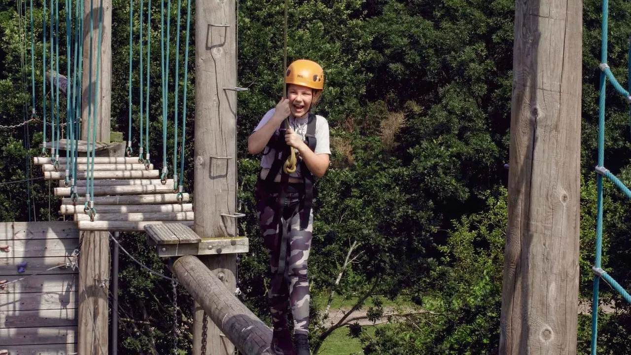 Young person wearing yellow helmet and harness on outdoor treetop adventure course with wooden platforms and ropes.