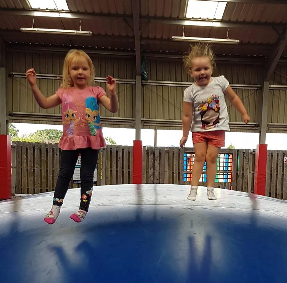 Two young children jumping on a blue trampoline in an indoor play facility with wooden fencing and corrugated roof.