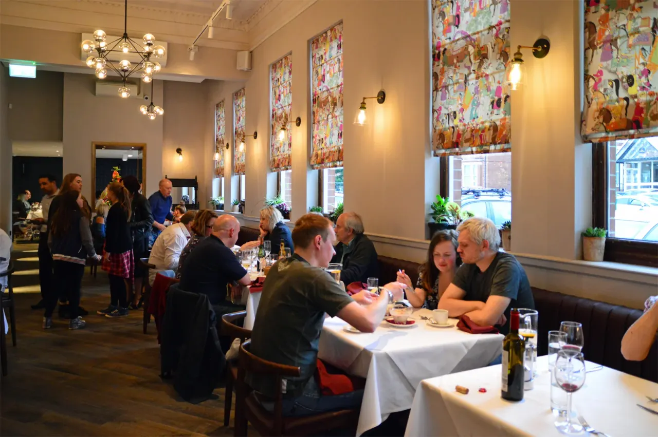 A restaurant interior with diners seated at tables with white tablecloths, colourful tapestries on walls, and pendant lighting overhead.