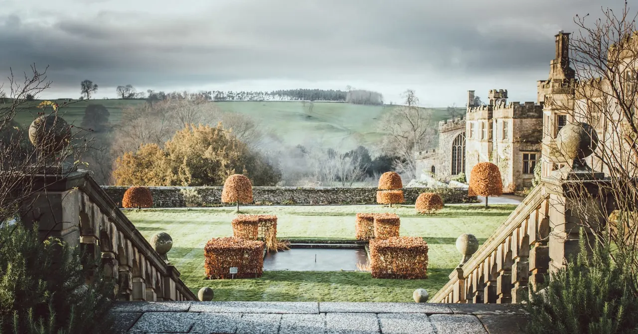 Stone castle with symmetrical formal gardens featuring ivy-covered topiary and a small pond, overlooking rolling countryside.