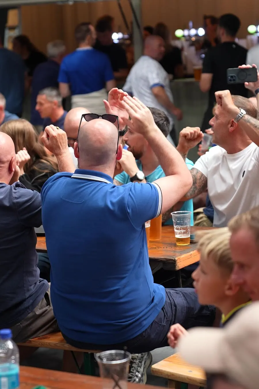 Crowded indoor event with people seated at tables with drinks, clapping and cheering, some recording on phones.