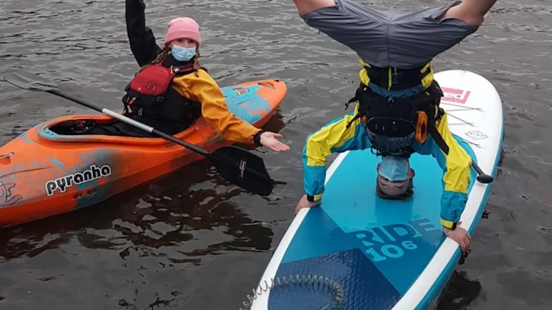 Two people in wetsuits and buoyancy aids on paddleboards in water, one holding a paddle.