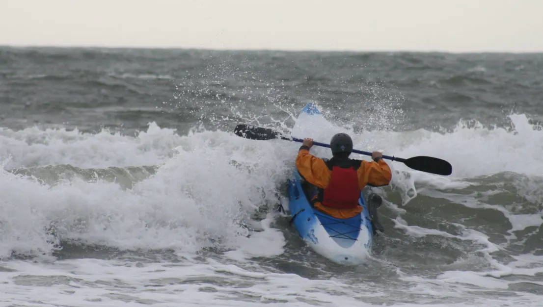 Person in orange jacket and blue kayak paddling through white-capped waves on rough sea.