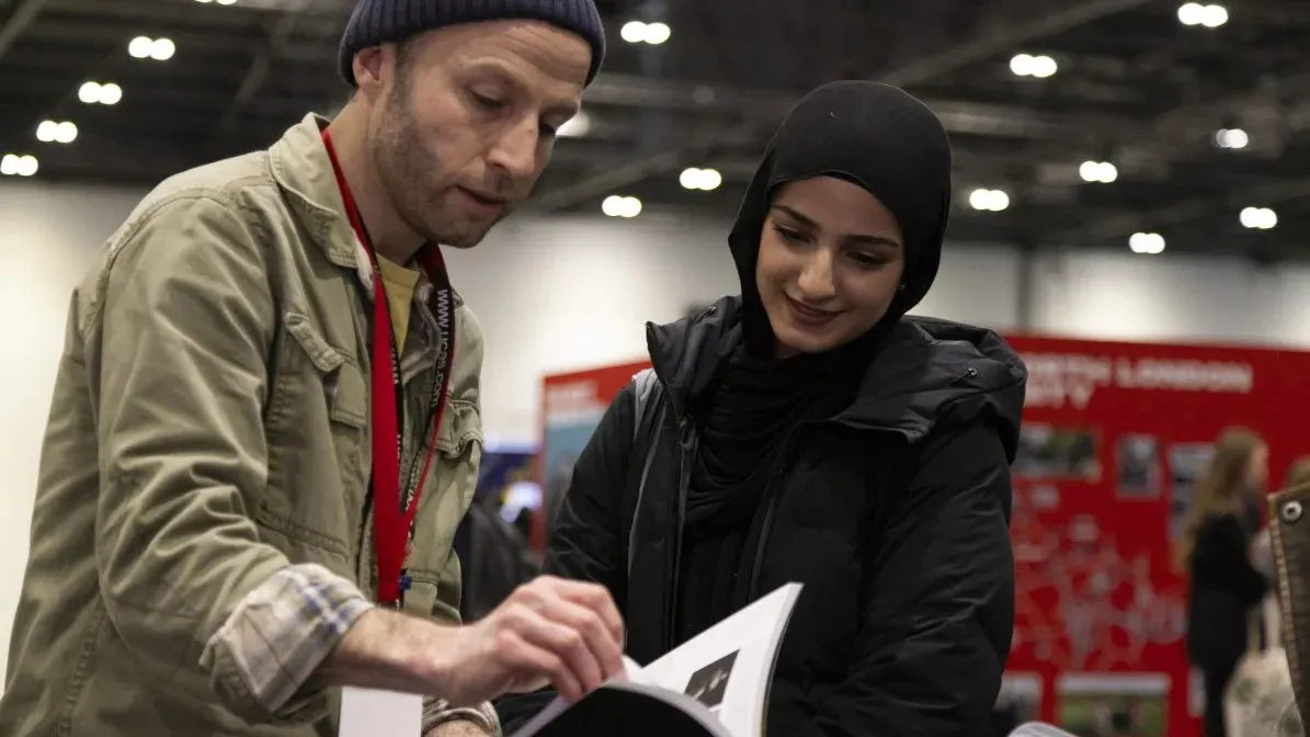 A staff member wearing a lanyard shows a visitor information on a leaflet inside an indoor venue with red signage and overhead lighting.