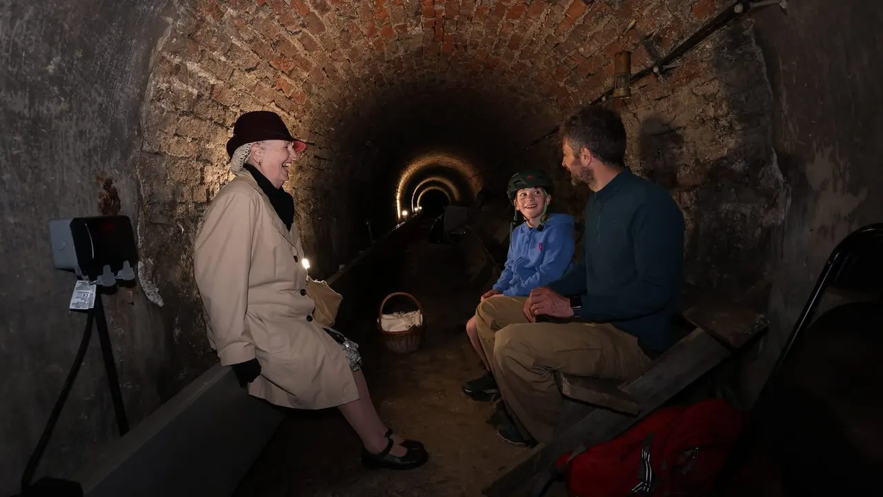 A guide in a bowler hat leads a group of visitors through a dimly lit brick tunnel with atmospheric lighting.