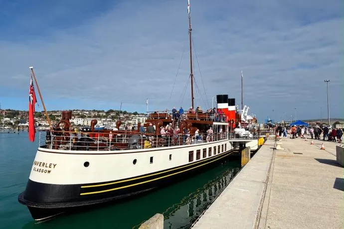 Vintage paddle steamer moored at a seaside pier with passengers boarding, coastal town visible beyond.