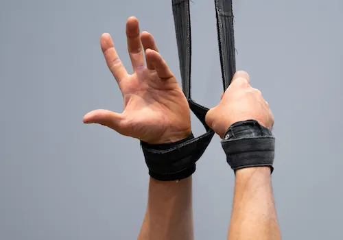 Person's hands raised upward while wearing black aerial straps against a clear sky.