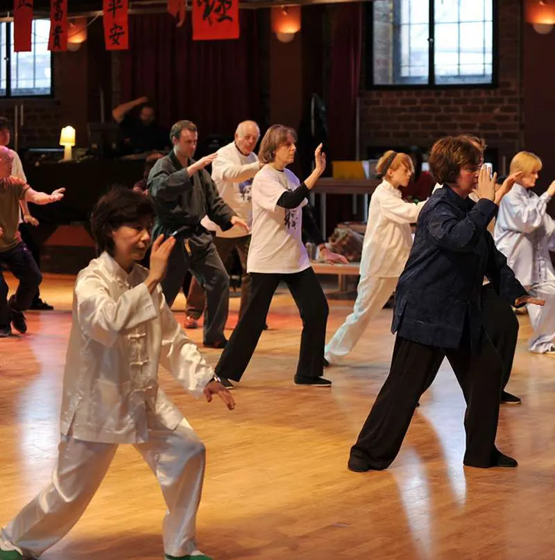 Group of adults and children practising tai chi together in a studio with wooden flooring and red lantern decorations.