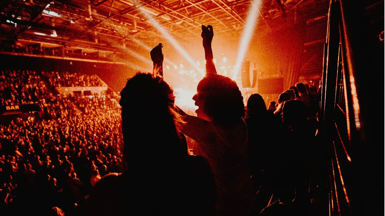High-angle view of a packed arena crowd and stage lights during a live concert, with a spectator in the foreground raising both arms.