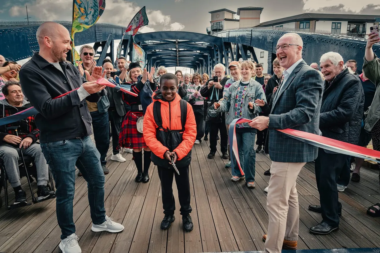 A child in an orange jacket stands on a wooden pier surrounded by adults cutting a red ribbon during an opening ceremony, with a heritage ship and crowd in the background.