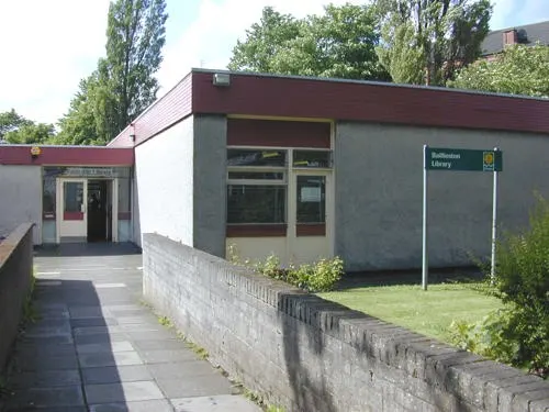 Modern single-storey library building with grey walls and burgundy roof, surrounded by trees and grass.