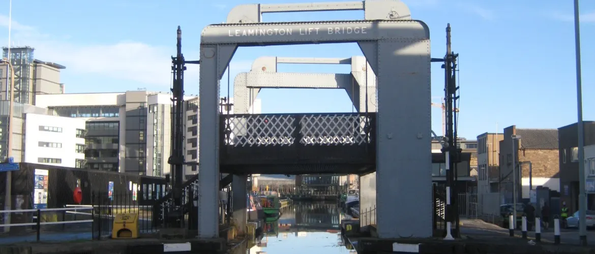 The Leamington Lift Bridge spanning a canal with a narrowboat moored below and urban buildings surrounding the waterway.