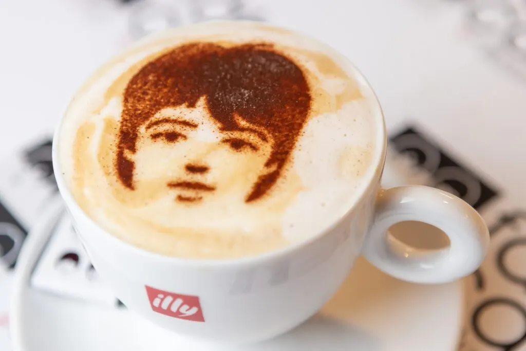 Cup of coffee with latte art portrait of a person with 1960s-style haircut.