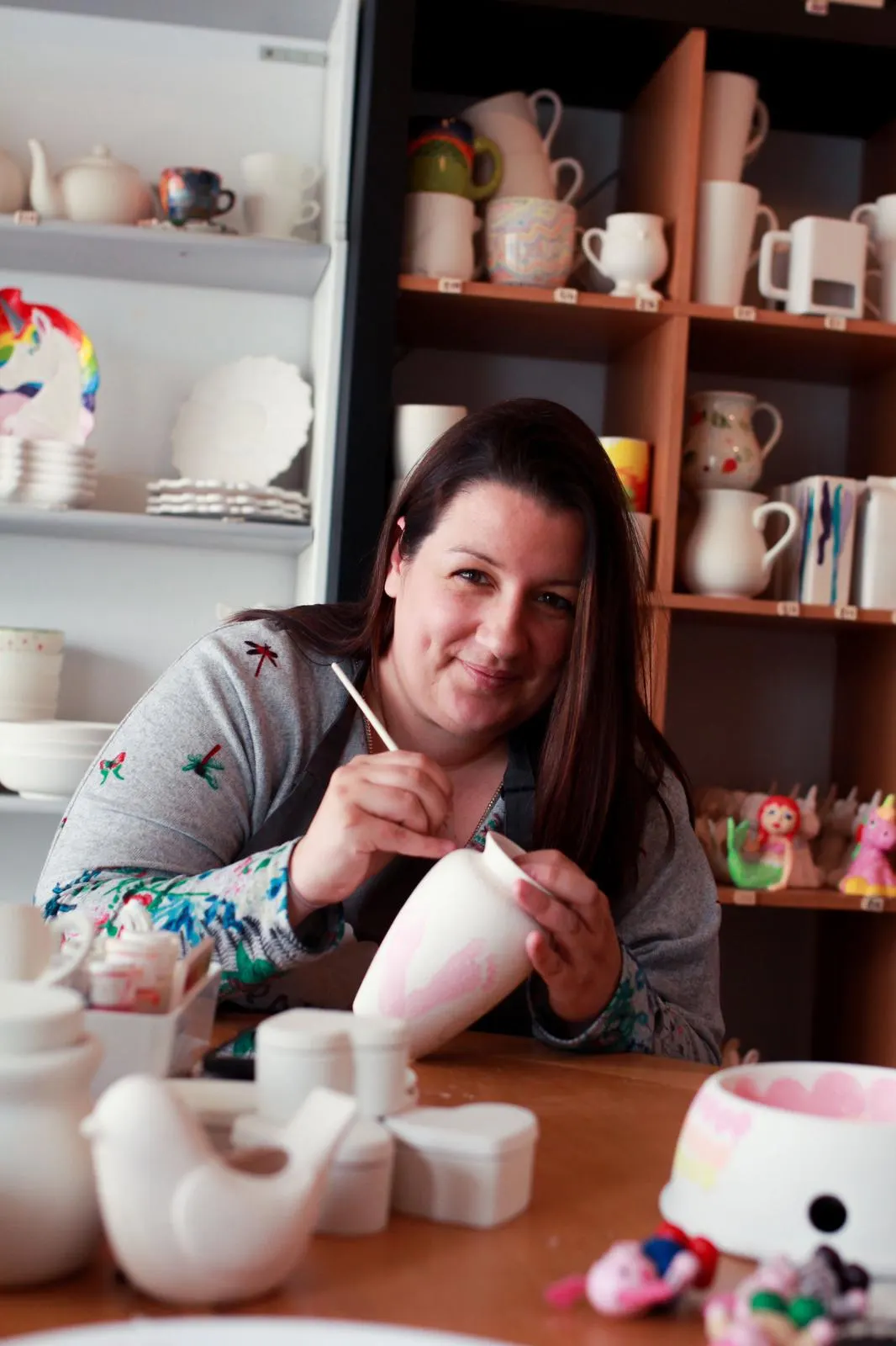 Woman painting a ceramic mug at a pottery studio table, with shelves of finished pottery and supplies behind her.