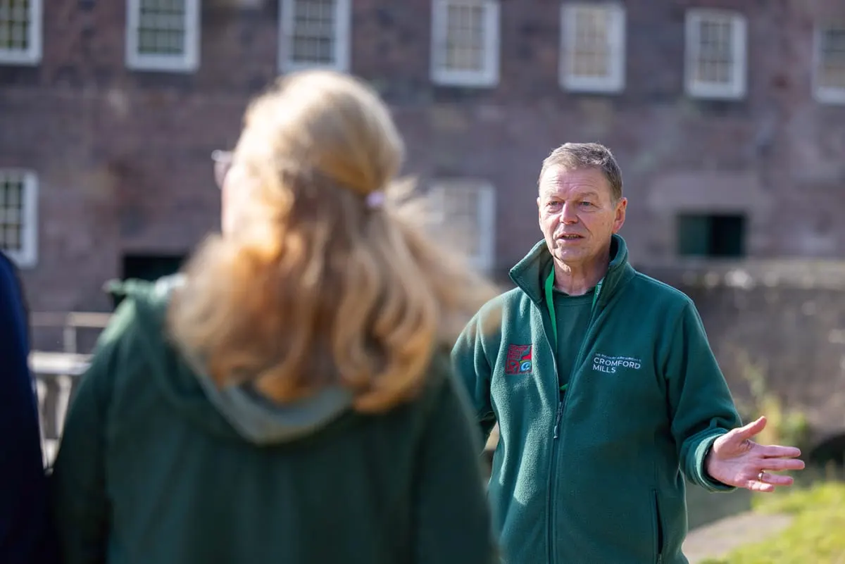 A man in a green Cromford Mills fleece speaks to a woman with blonde hair, with a historic brick building behind them.
