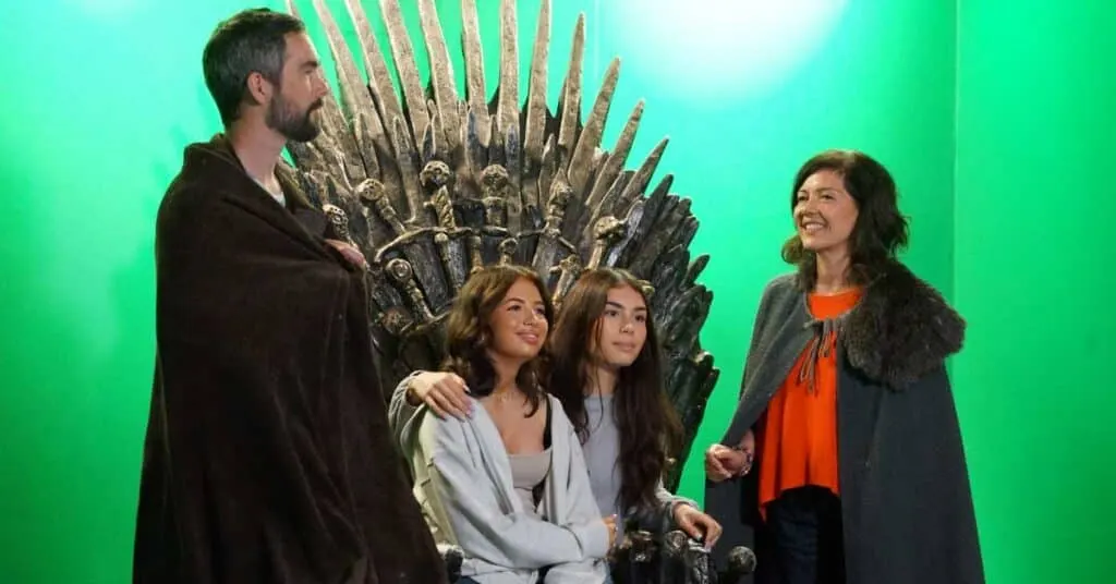 Four visitors posing together in front of a large ornate iron throne against a bright green screen.