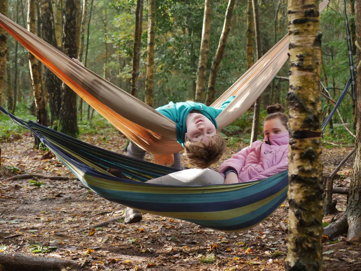 Two children relaxing in a hammock strung between trees in a woodland setting.