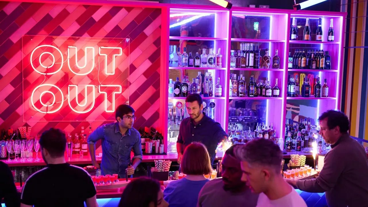 Bartenders serve drinks to customers at a neon-lit bar with illuminated spirit bottles on shelves behind them.