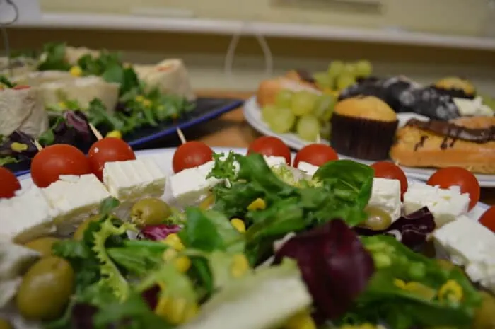 Platters of catering food including salads, cheese, tomatoes and greens arranged on tables.