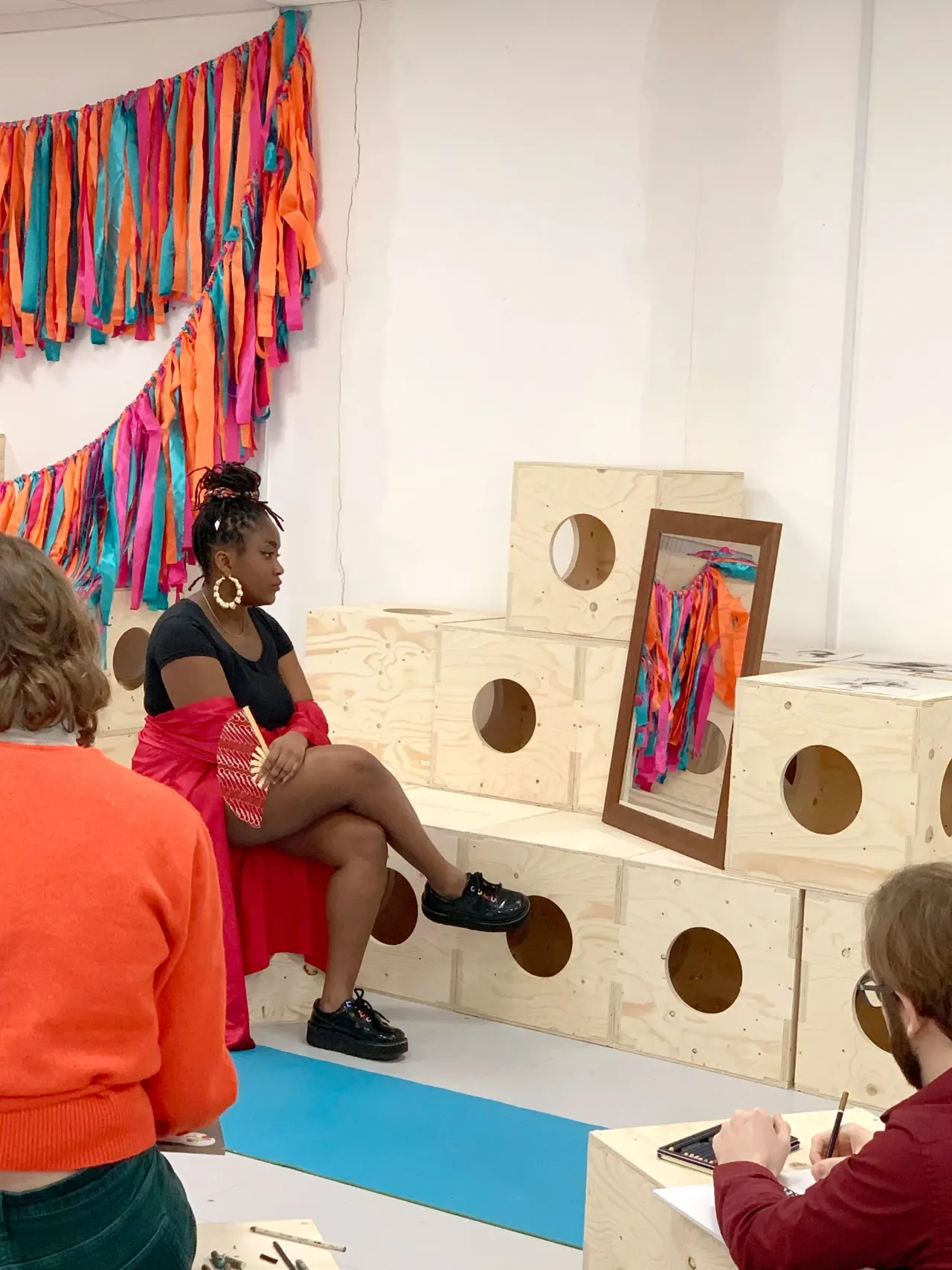 Young woman sitting on wooden play structures with colourful fabric strips, whilst others observe in an interactive art installation.