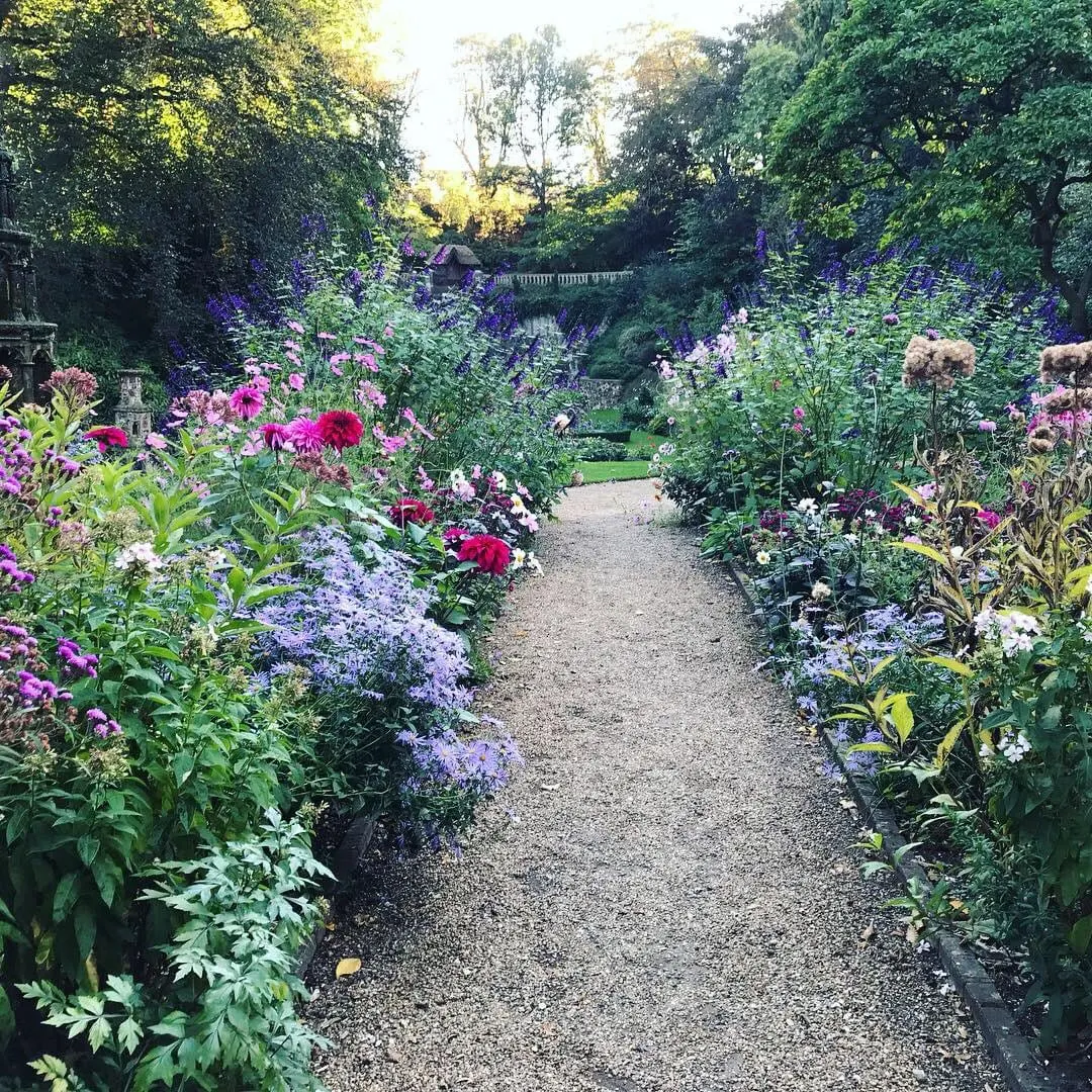 Gravel pathway through a formal garden lined with pink, purple and red flowering plants, with mature trees and a stone balustrade visible in the distance.
