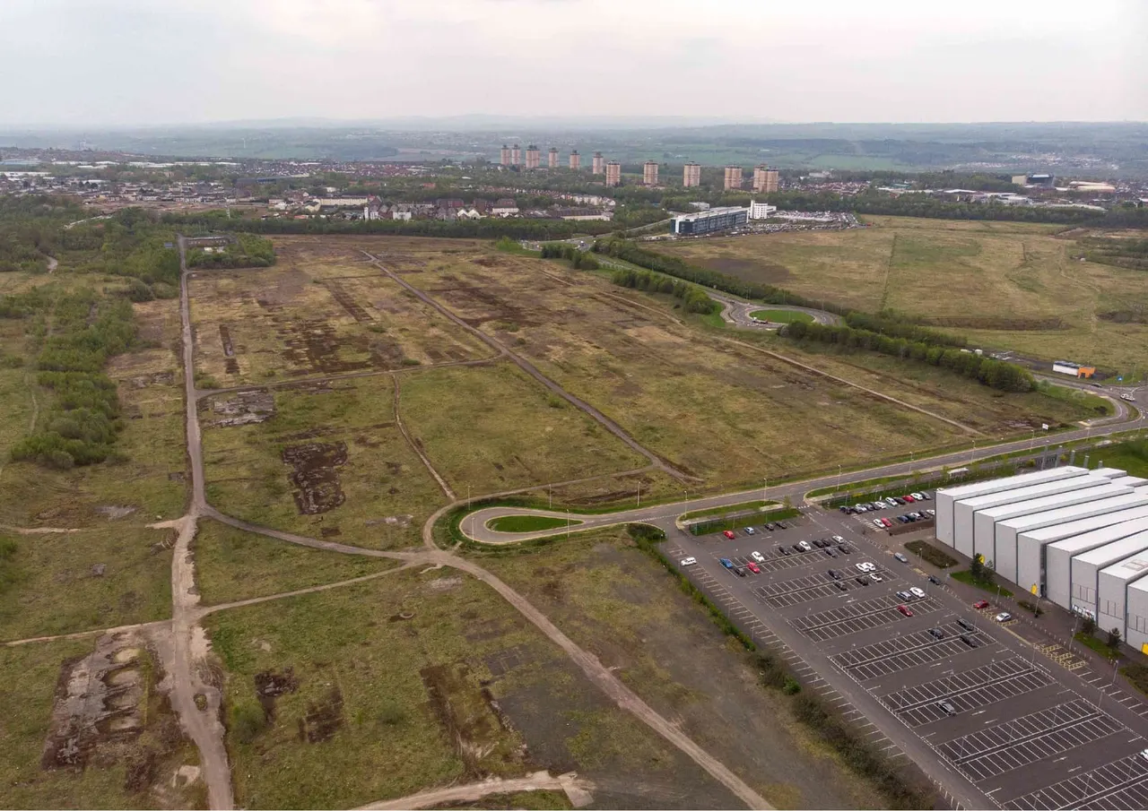 Aerial view of parkland with fields, a car park, warehouse buildings and distant urban landscape.