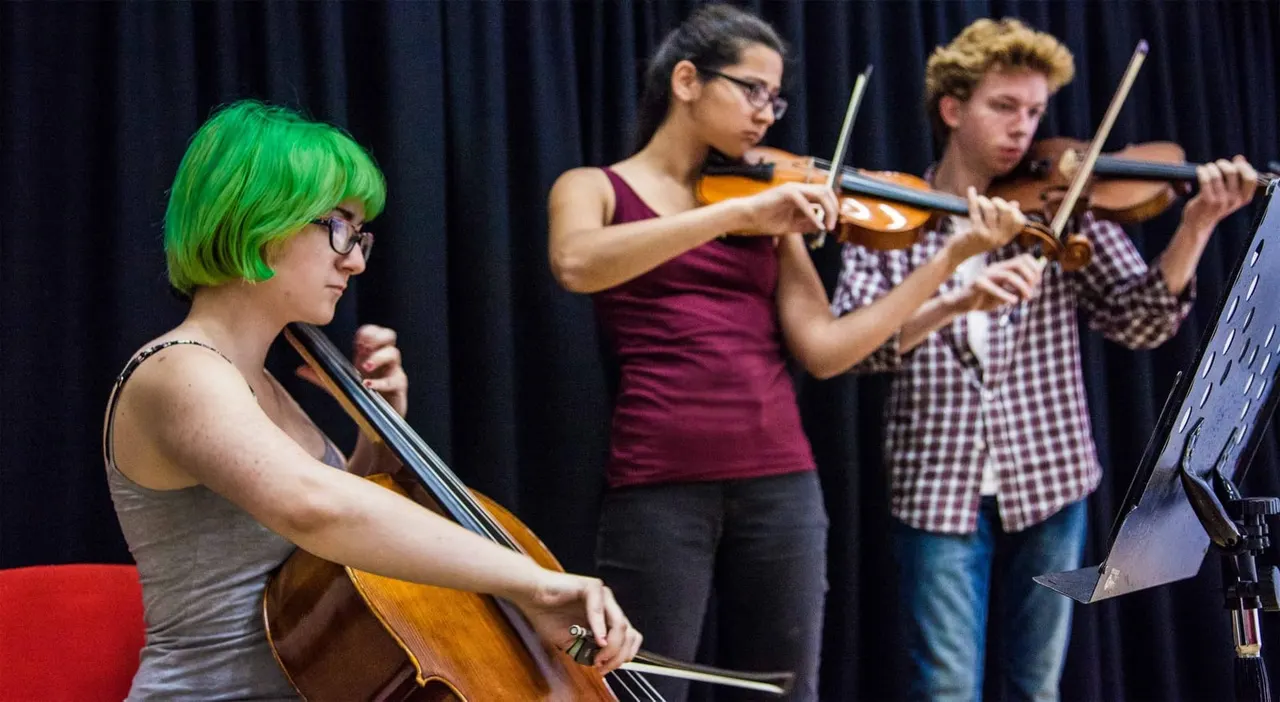 Young musicians performing together on stage, playing violin and cello against a dark blue curtain backdrop.