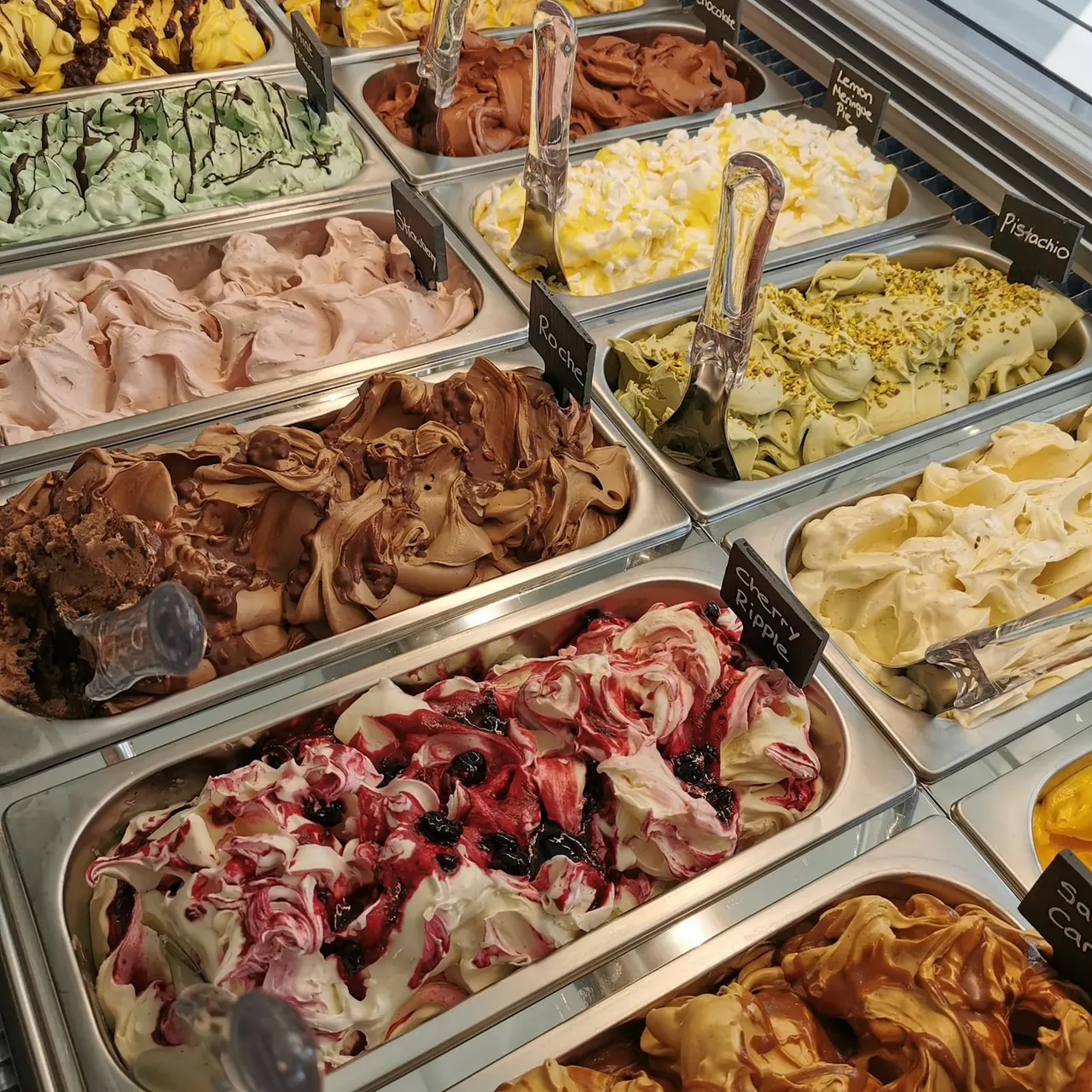 Display case of various ice cream flavours including chocolate, pistachio, berry ripple and mint in metal tubs with serving scoops.