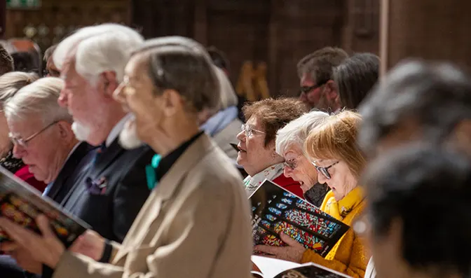 Visitors of mixed ages gathered indoors holding service sheets and hymn books.