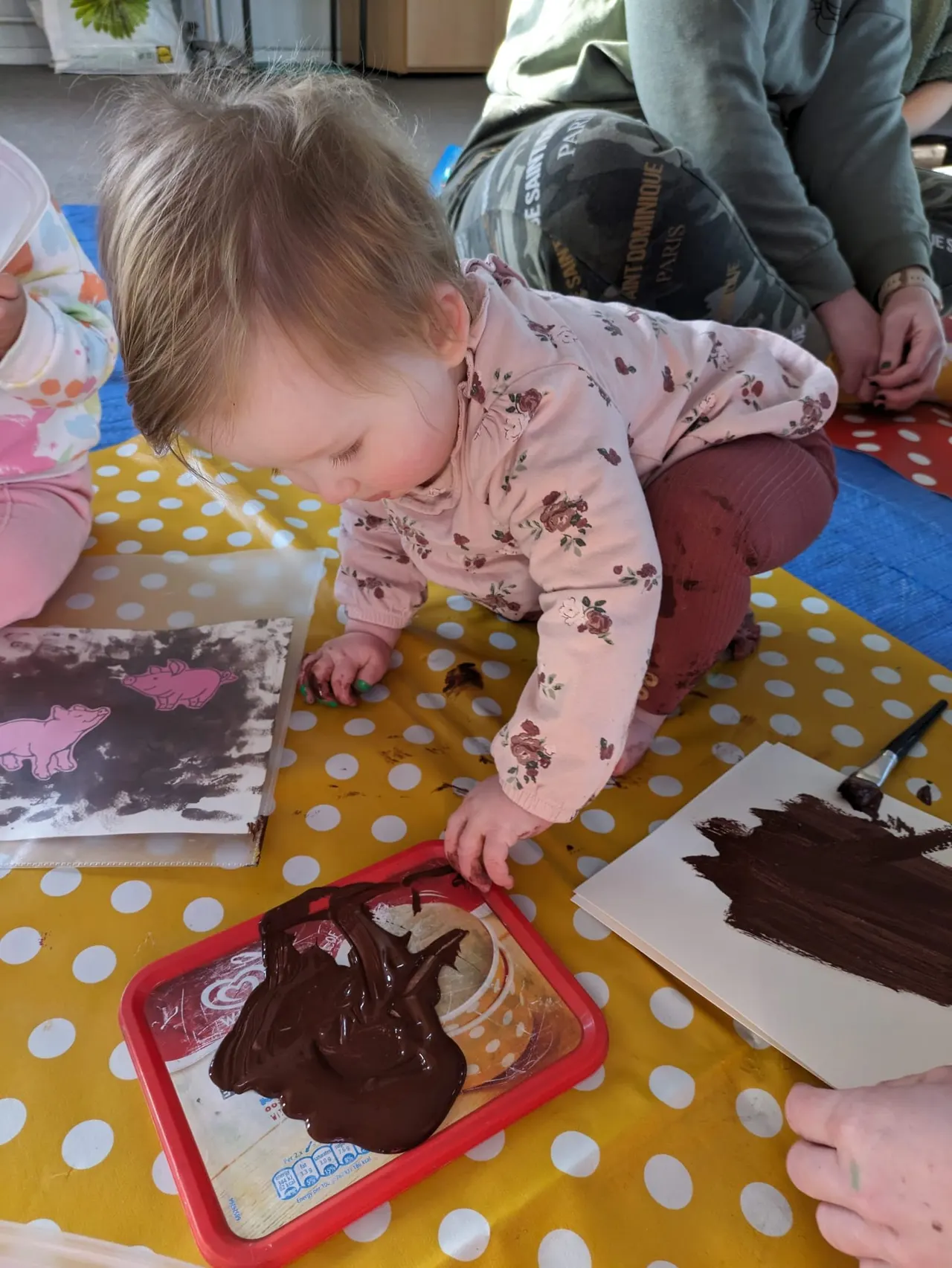 Toddler in pink floral clothing painting with brown paint on a yellow polka-dot mat during a supervised craft activity.
