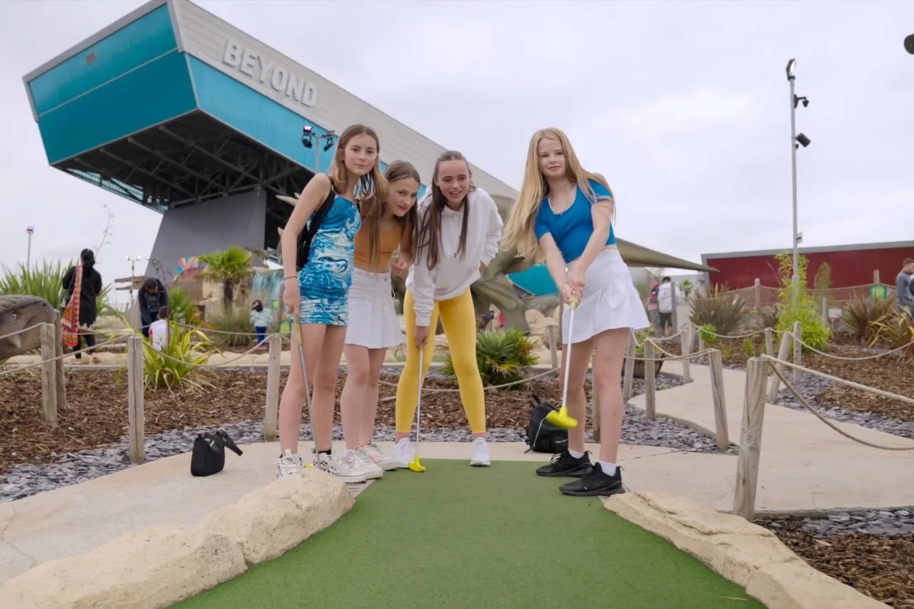 Four young people playing mini golf on an artificial green course with a large blue building in the background.