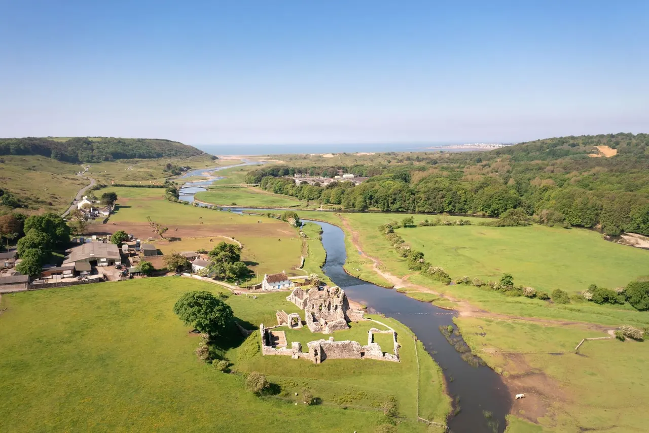 Aerial view of ruined castle beside a meandering river in a lush valley with green fields, woodlands and coastal views in the distance.