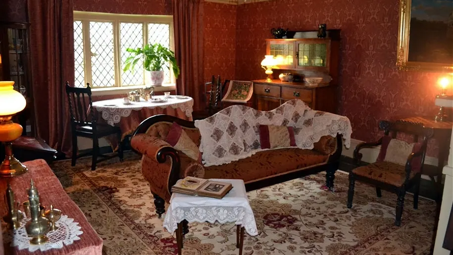 Victorian sitting room with burgundy wallpaper, period furniture including a leather sofa and wooden chairs, small dining table by the window, and decorative accessories throughout.