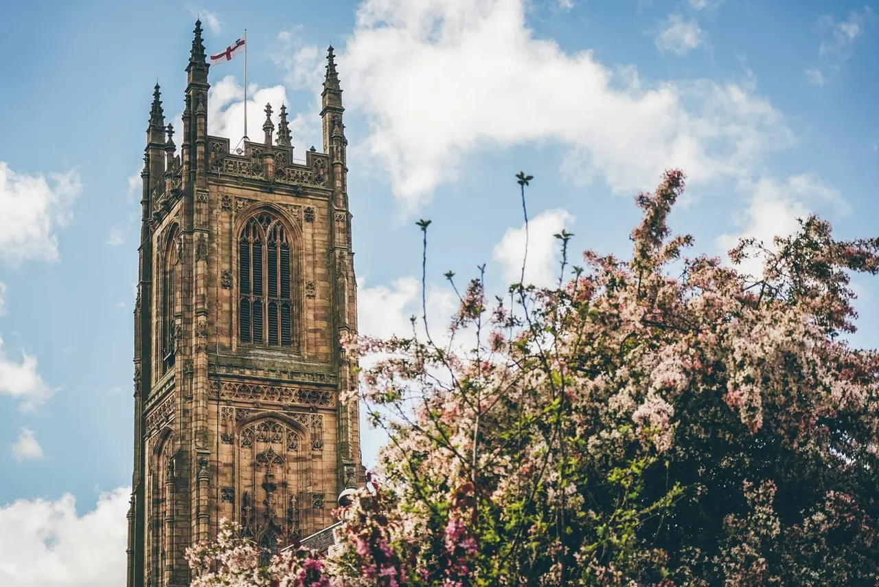 Gothic stone cathedral tower with ornate spires and Canadian flag, framed by flowering shrubs and blue sky with white clouds.