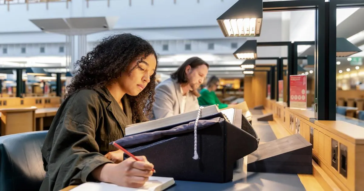 Young person studying at a table in a library reading room with other visitors in the background.