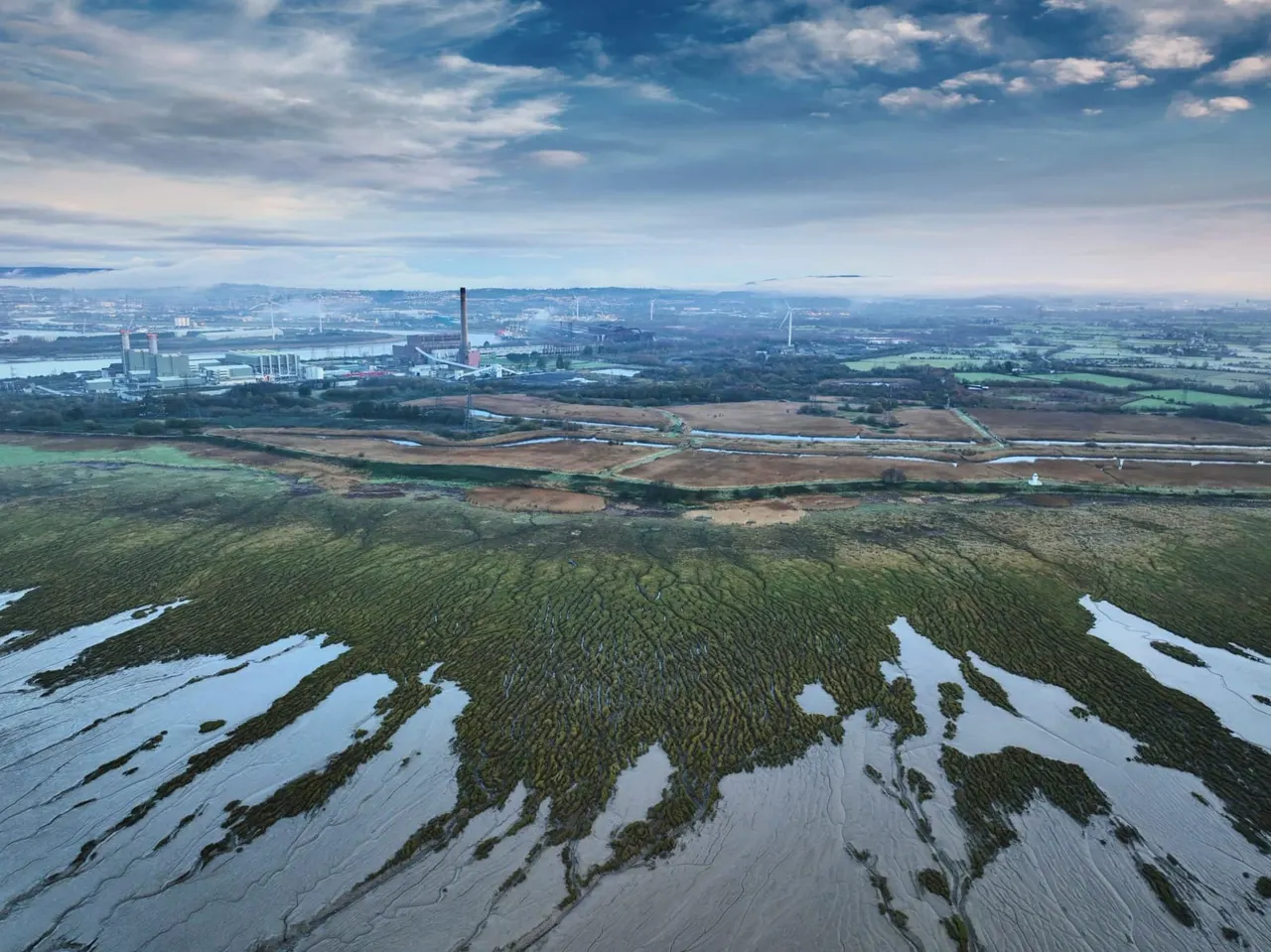 Aerial view of wetland marshes with shallow water and vegetation, industrial complex and distant cityscape beyond.