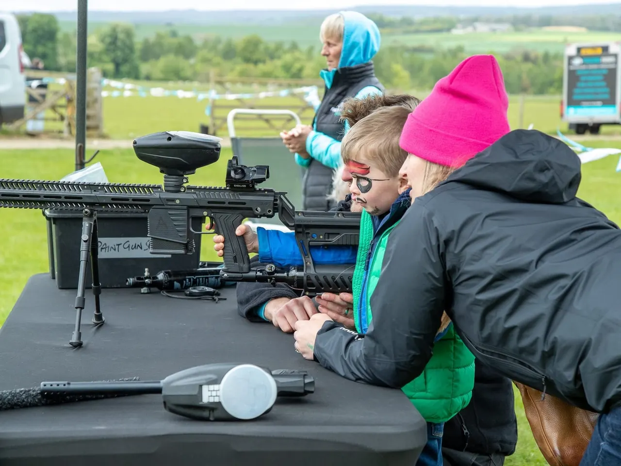 Children handling paintball guns at an outdoor leisure activity, with countryside and spectators visible beyond.