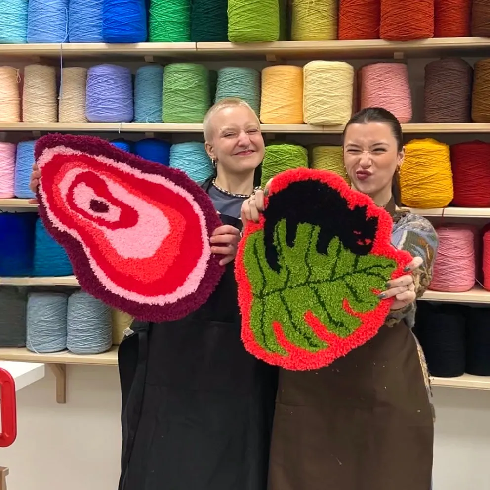 Two people hold handmade tufted textile pieces with spiral and abstract designs in front of shelves of coloured yarn.
