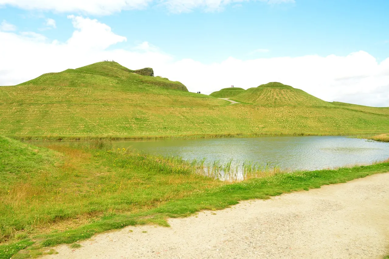 Green grass mounds rise beside a still pond under blue sky, with a gravel path in the foreground.